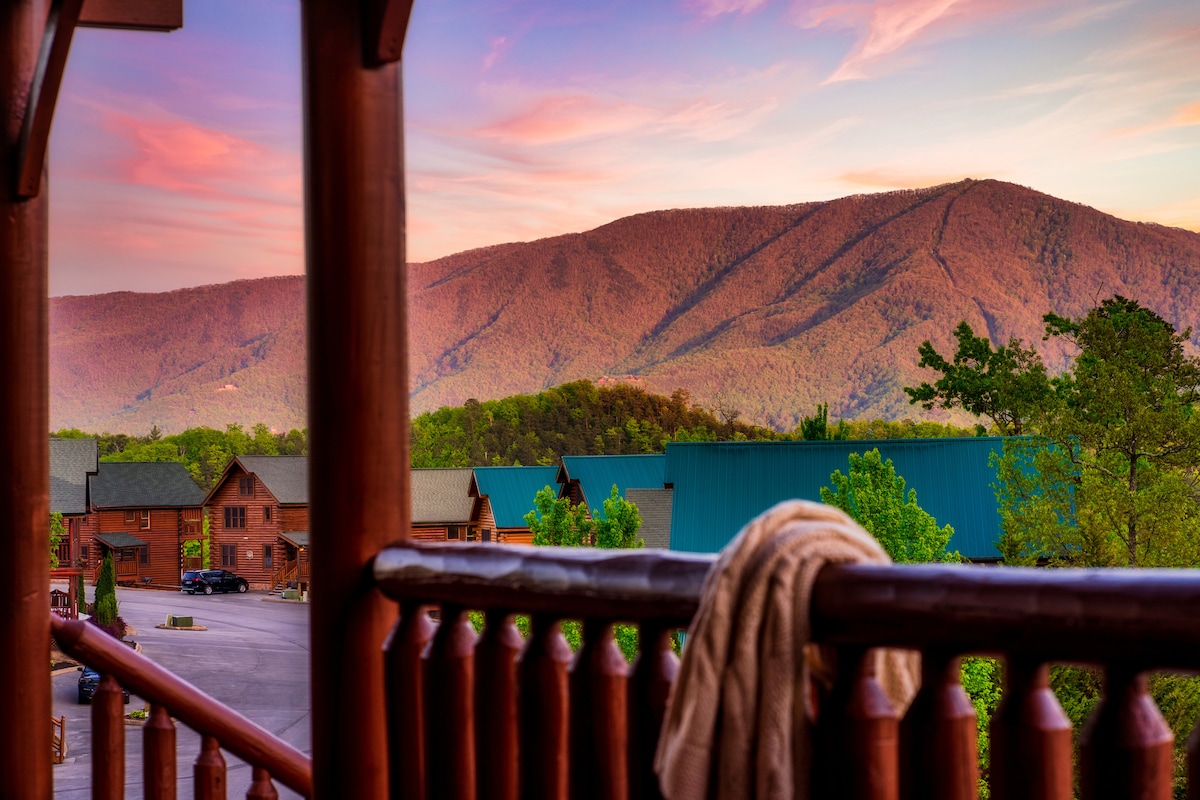 A balcony view reveals rolling hills and mountain peaks bathed in soft sunset hues. Wooden railing frames the scene, while a seating area is visible nearby. Nearby cabins and a parking area are set against the vibrant landscape.