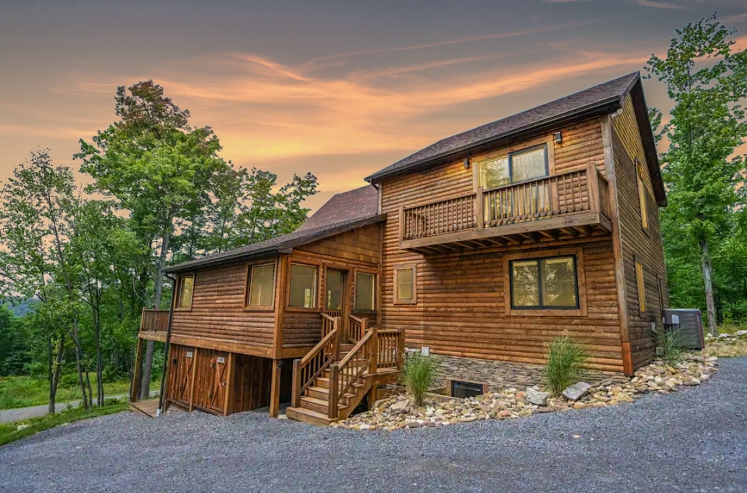 A wooden house is set against a sunset sky, surrounded by trees. Steps lead to the entrance, and multiple balconies showcase expansive views, creating a blend of indoor and outdoor living. Natural stone accents the landscaping, complementing the rustic charm of the property.