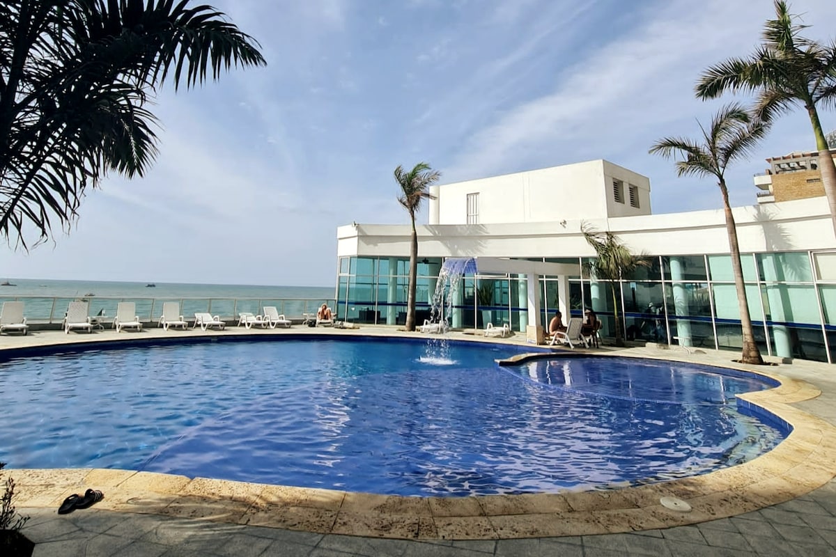 A large outdoor swimming pool is showcased with inviting blue water, surrounded by palm trees. Sun loungers are positioned around the pool area, with a beachfront view visible through expansive glass windows of the building.