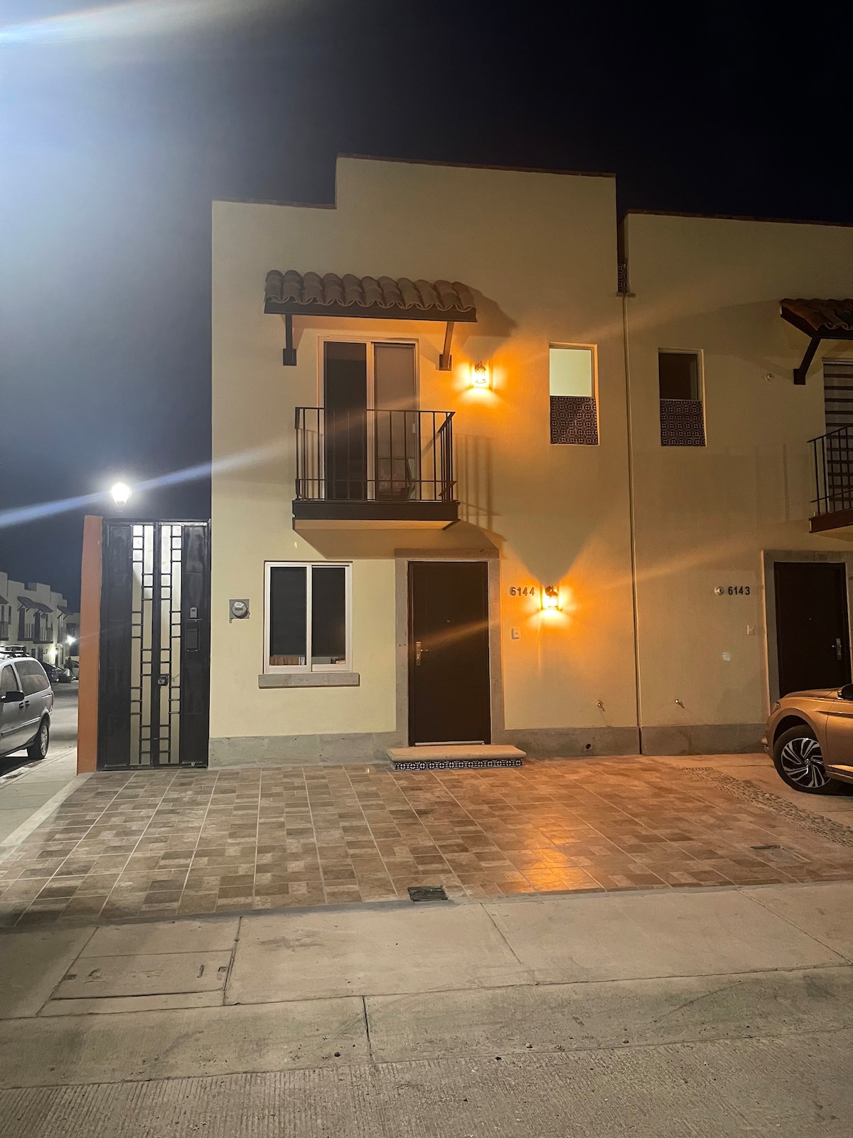 A modern two-story home is illuminated by warm exterior lights, featuring a front door flanked by windows. A balcony is visible on the second level. Paved stone pathways lead to the entrance, with vehicle parking available in front of the property.