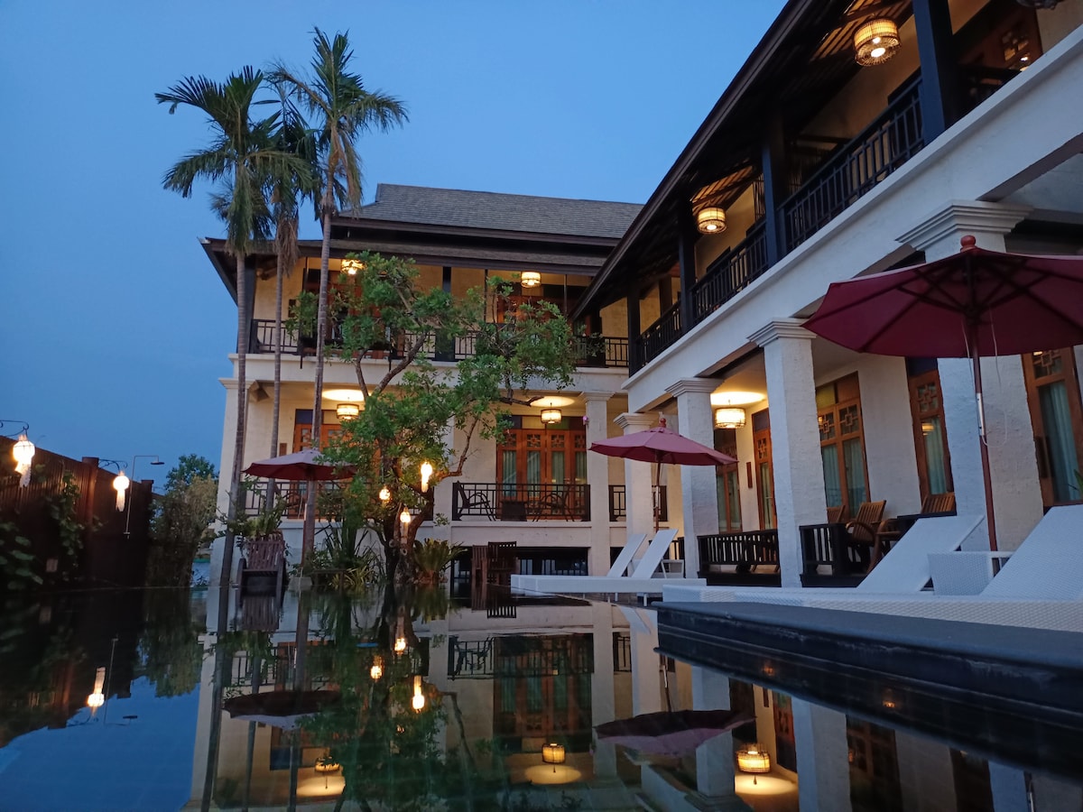 The hotel exterior is shown at dusk, featuring a three-story structure with balconies. Poolside lounge chairs are visible, accompanied by red umbrellas. Lush greenery is present, and soft lighting reflects off the surface of the nearby saltwater pool, enhancing the tranquil atmosphere.