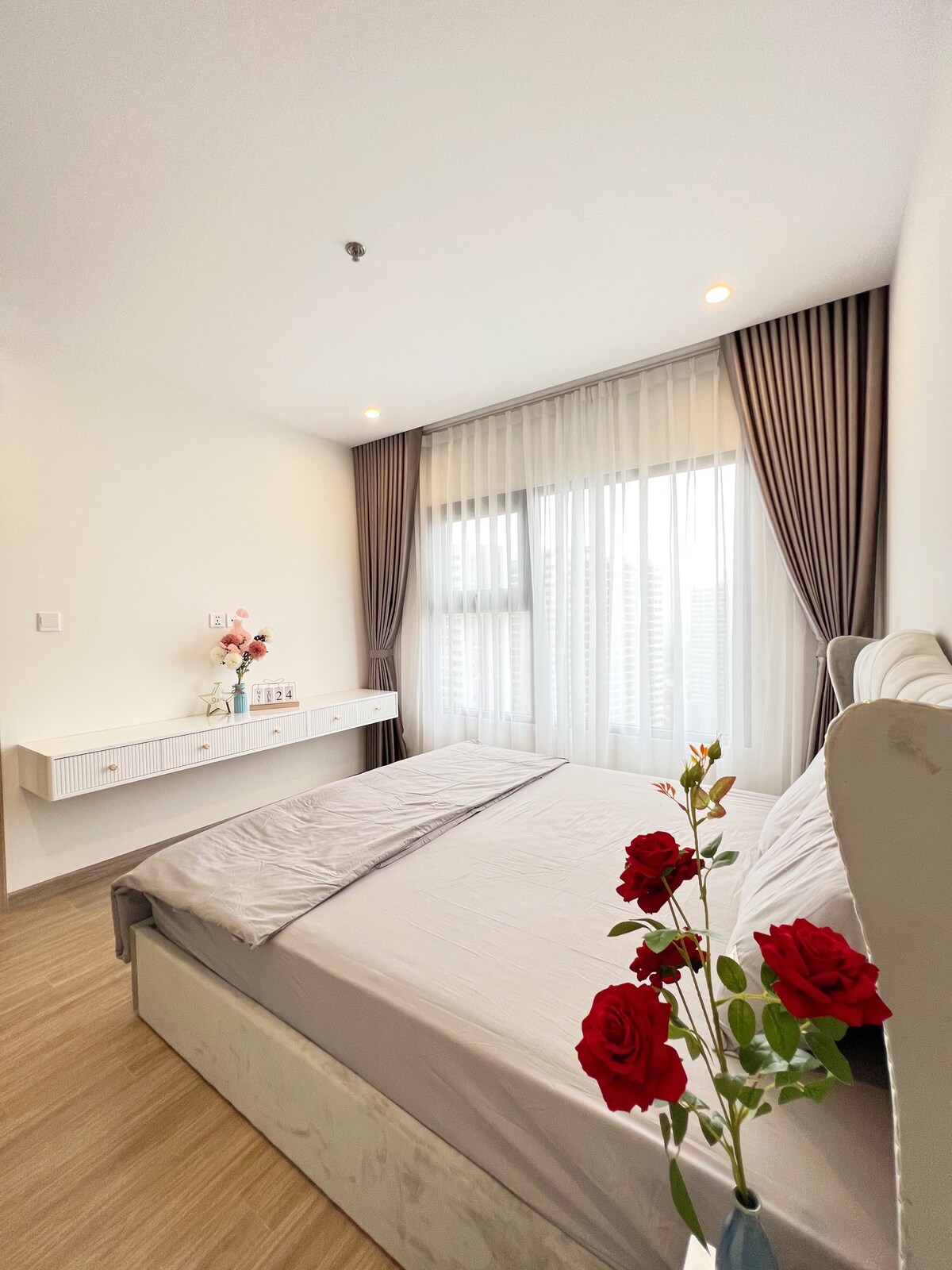 A bright bedroom features a spacious bed with a light-colored comforter. A wall-mounted shelf displays decorative items. Sheer curtains allow natural light to filter through large windows, and a vase of red roses adds a touch of color to the serene space.