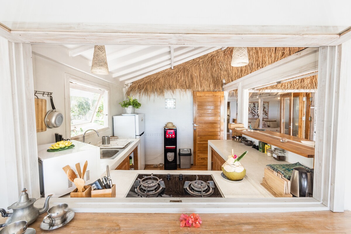 The kitchen area features a spacious layout with natural materials and ample counter space. A stove with a sleek surface is positioned centrally, with organized utensils and fresh fruit displayed. Bright light enters through windows, creating an inviting cooking space with rustic design elements.