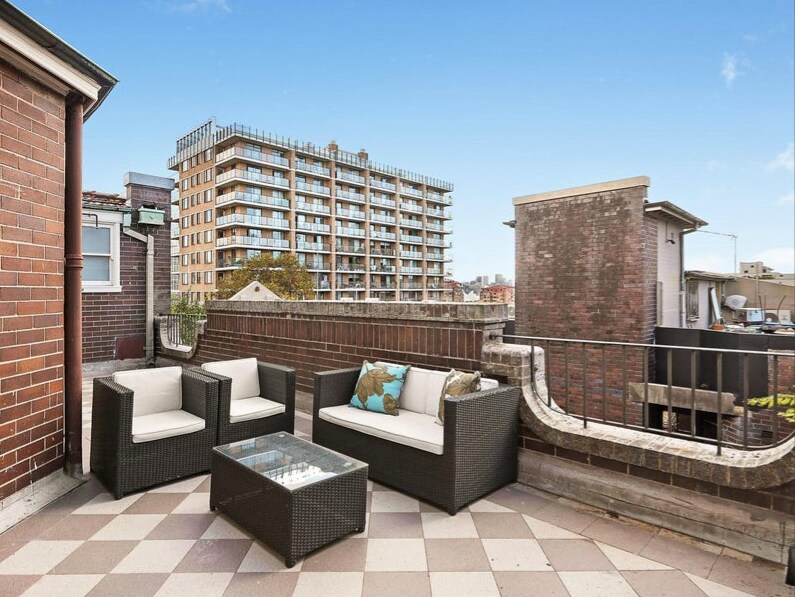 A shared rooftop area is presented, featuring a modern seating arrangement with a glass table and plush chairs. The space is bordered by brick walls and overlooks the surrounding buildings, providing glimpses of the city skyline under a clear blue sky.