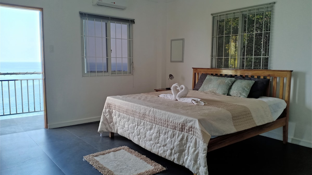 A spacious bedroom features a large bed with a neutral-colored quilt and decorative pillows. Soft light enters through the nearby window, which offers a view of the ocean. A small rug lies at the foot of the bed, and a towel arrangement adds a touch of comfort.