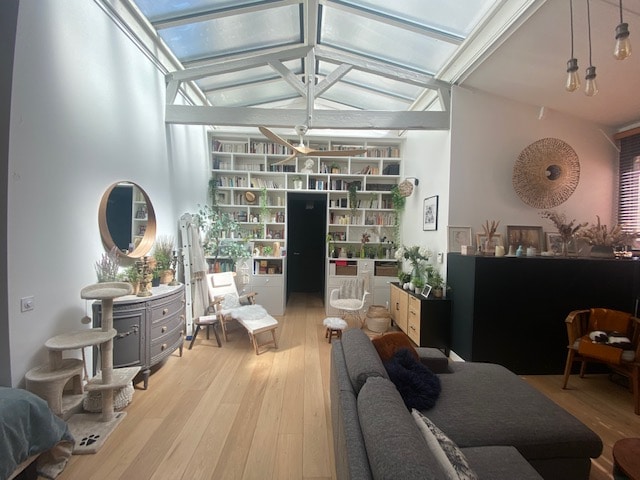 A spacious living area is featured, highlighted by a glass roof that allows ample natural light. A collection of books and decorative items is displayed on shelves, while various seating options, including two stylish chairs and a gray sofa, create inviting spaces for relaxation.