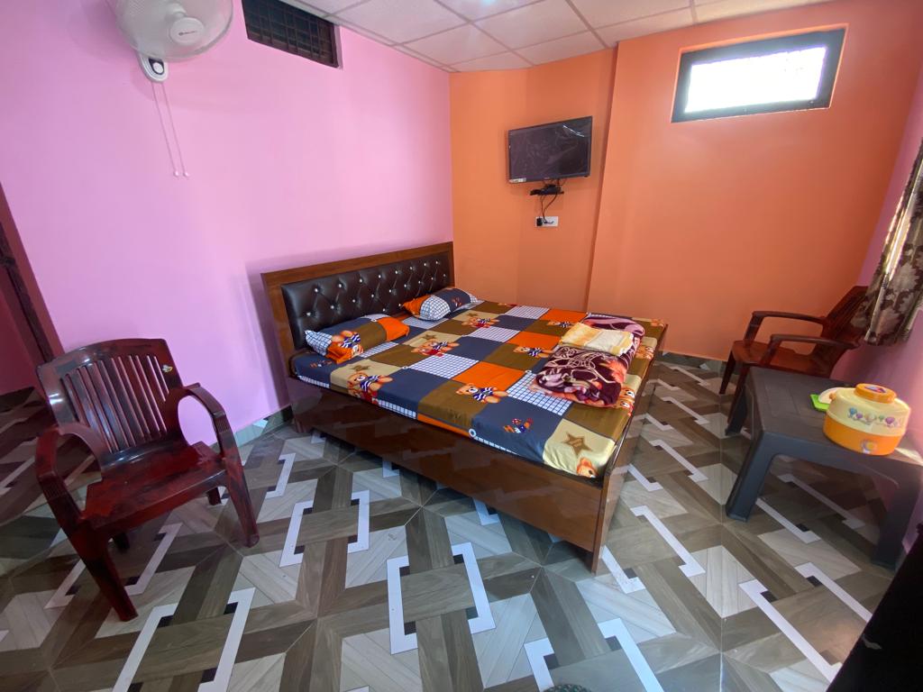 A cozy room features a spacious bed with a patterned blanket. Two wooden chairs are present, alongside a small table. A flat-screen TV is mounted on the wall, with natural light filtering through a window above the bed. The floor displays a unique geometric design.