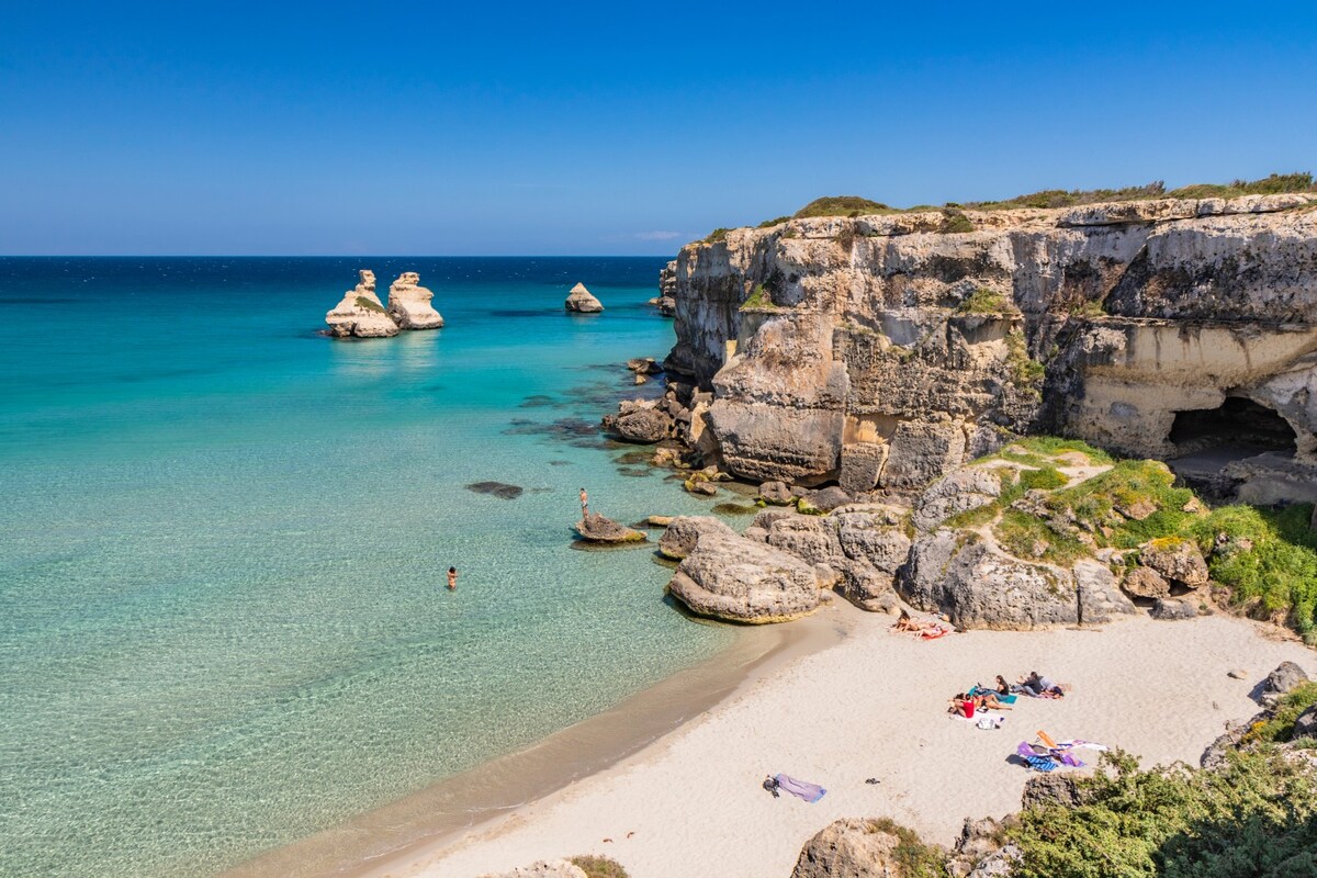 A serene coastline is depicted, featuring golden sandy shores and clear turquoise waters. Natural rock formations rise from the sea, with a small beach area where towels and beachgoers are present. The backdrop showcases cliffs adorned with lush greenery under a bright blue sky.