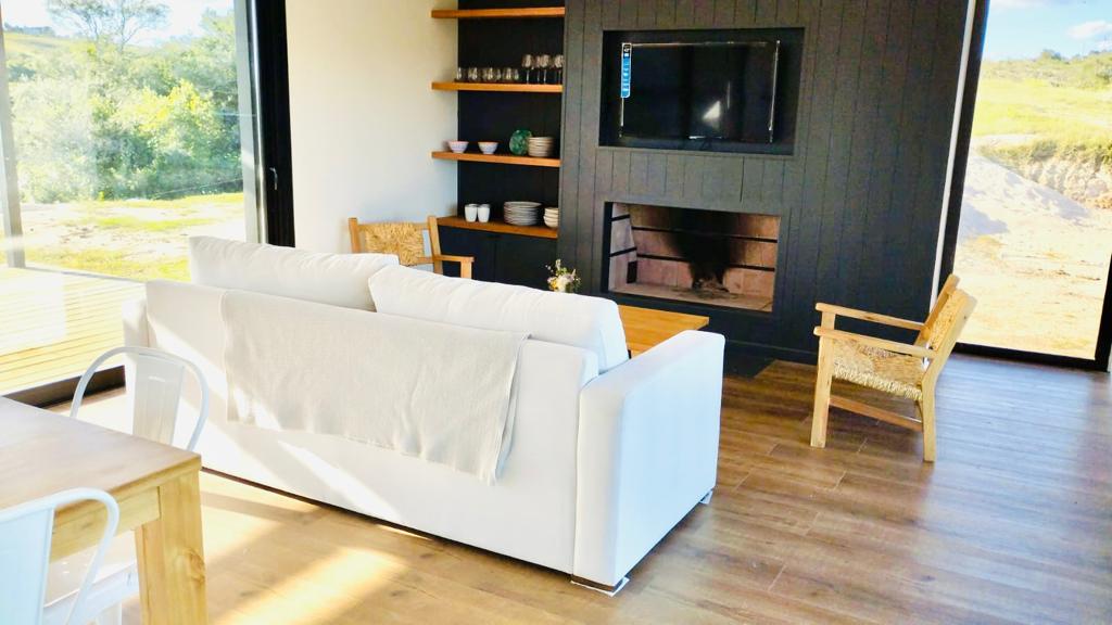 A modern living area features a white sofa with a light throw blanket, accented by natural wood shelving displaying dishware. A flat-screen television is mounted above a cozy fireplace, while a wicker chair complements the space. Large windows invite views of the surrounding landscape.