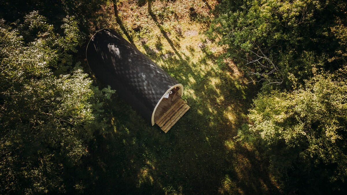 A camping pod is positioned amidst lush greenery, surrounded by trees. The pod features a rounded design with a wooden deck area extending from the entrance, blending harmoniously with the natural landscape. The dappled sunlight filters through the overhead leaves, casting soft shadows on the ground.