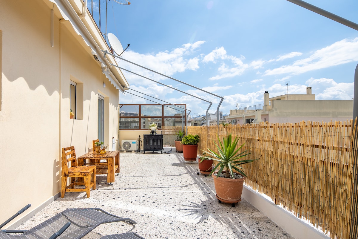 A spacious balcony is shown, furnished with wooden seating and a small table. Potted plants add greenery, and a sunshade extends overhead. The view includes the surrounding cityscape under a clear blue sky, with sunlight creating soft shadows along the floor.