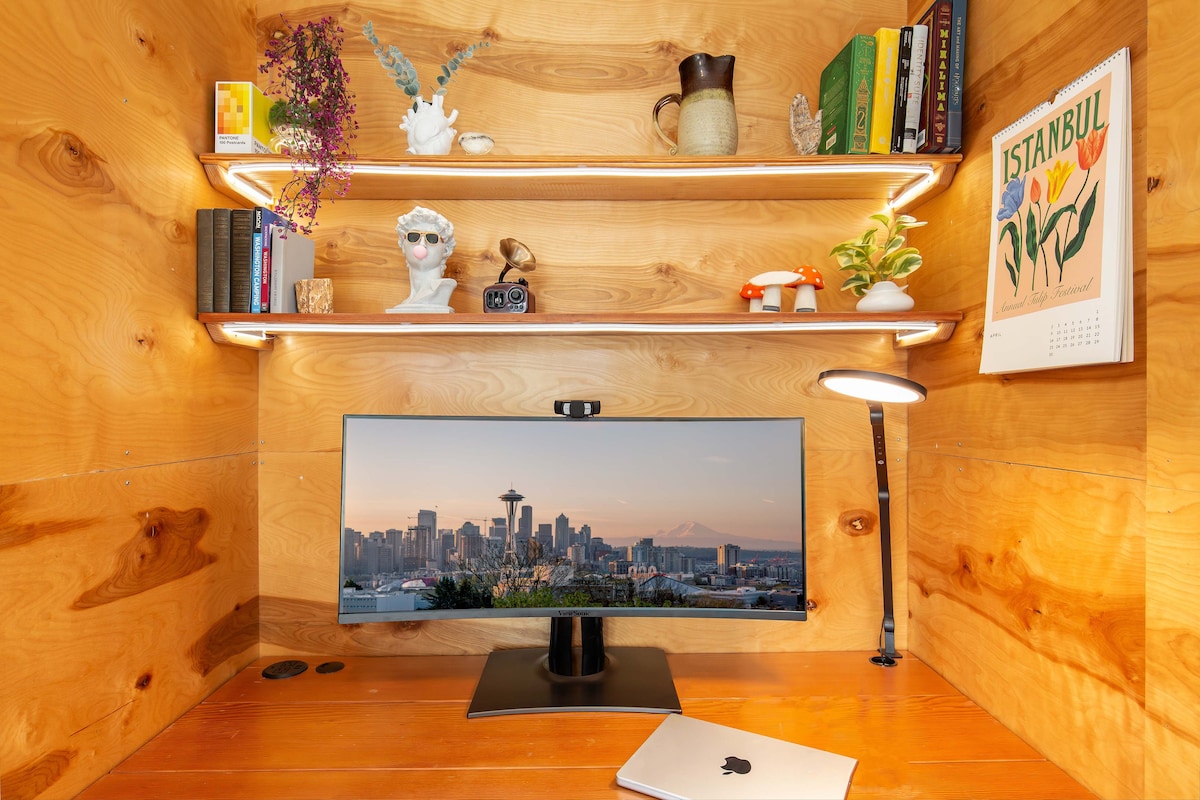 A compact workspace features a monitor displaying a city skyline. Floating shelves above hold books, a decorative bust, and plants. A sleek lamp provides illumination, while a laptop rests on the wooden surface. Warm wood paneling creates a cozy atmosphere.
