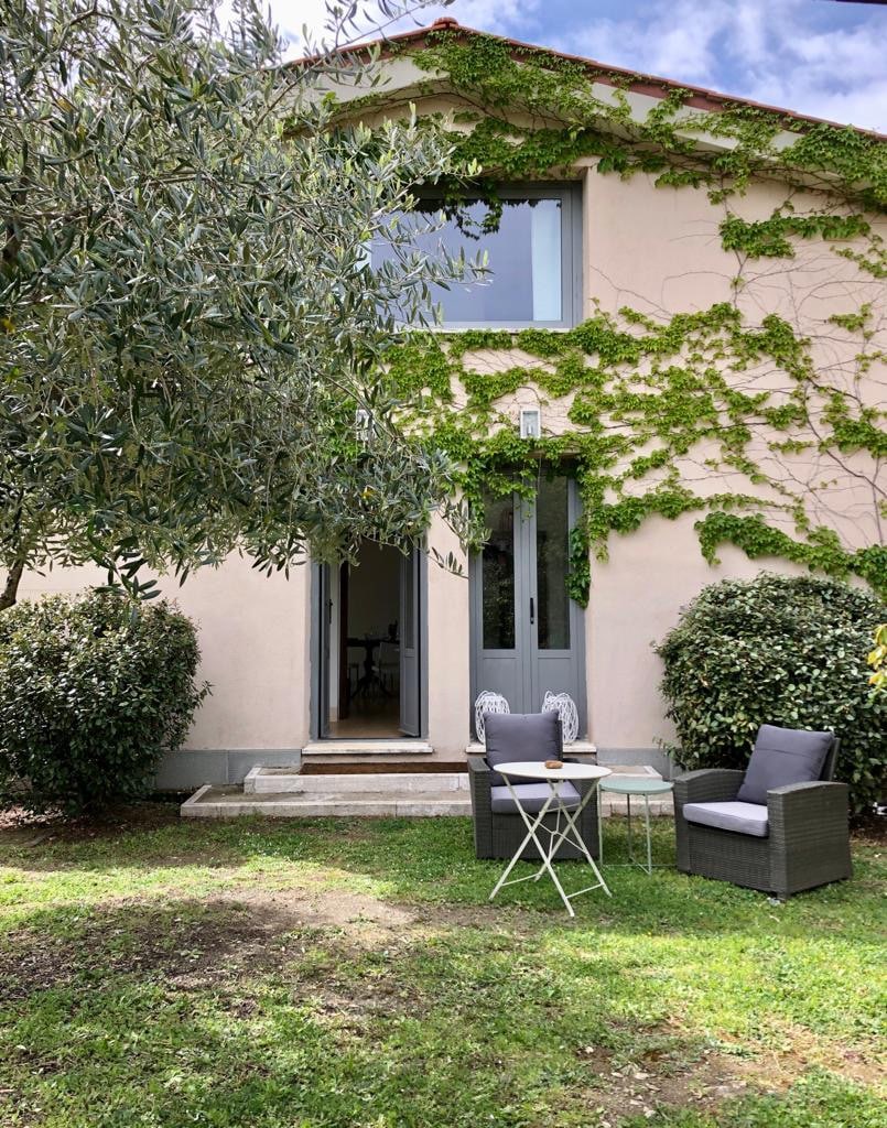 An inviting exterior of the independent apartment is showcased, featuring a light-colored facade adorned with climbing greenery. A small seating area with two chairs and a table is set on the grassy lawn, providing a welcoming space for relaxation.