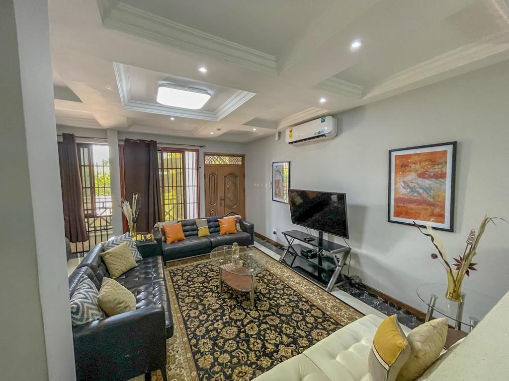 A spacious living room features a dark leather sectional sofa arranged around a glass coffee table. A large television is mounted on the wall opposite, with artfully arranged decorative items nearby. Bright natural light is filtered through large windows, enhancing the cozy atmosphere.