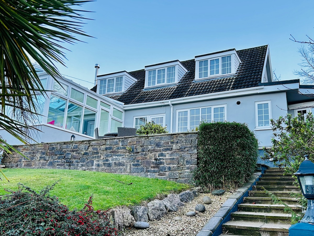 The exterior of a modern home is showcased, featuring a well-maintained stone wall and stairs leading up to a grassy yard. Large windows reflect natural light, with a conservatory visible on the side, framed by shrubs and palm plants.
