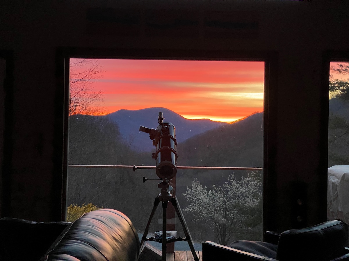 A stunning sunset paints the sky in vibrant hues behind a telescope, positioned in front of large windows. The silhouette of nearby mountains is visible against the colorful sky, creating a serene view from the living space.