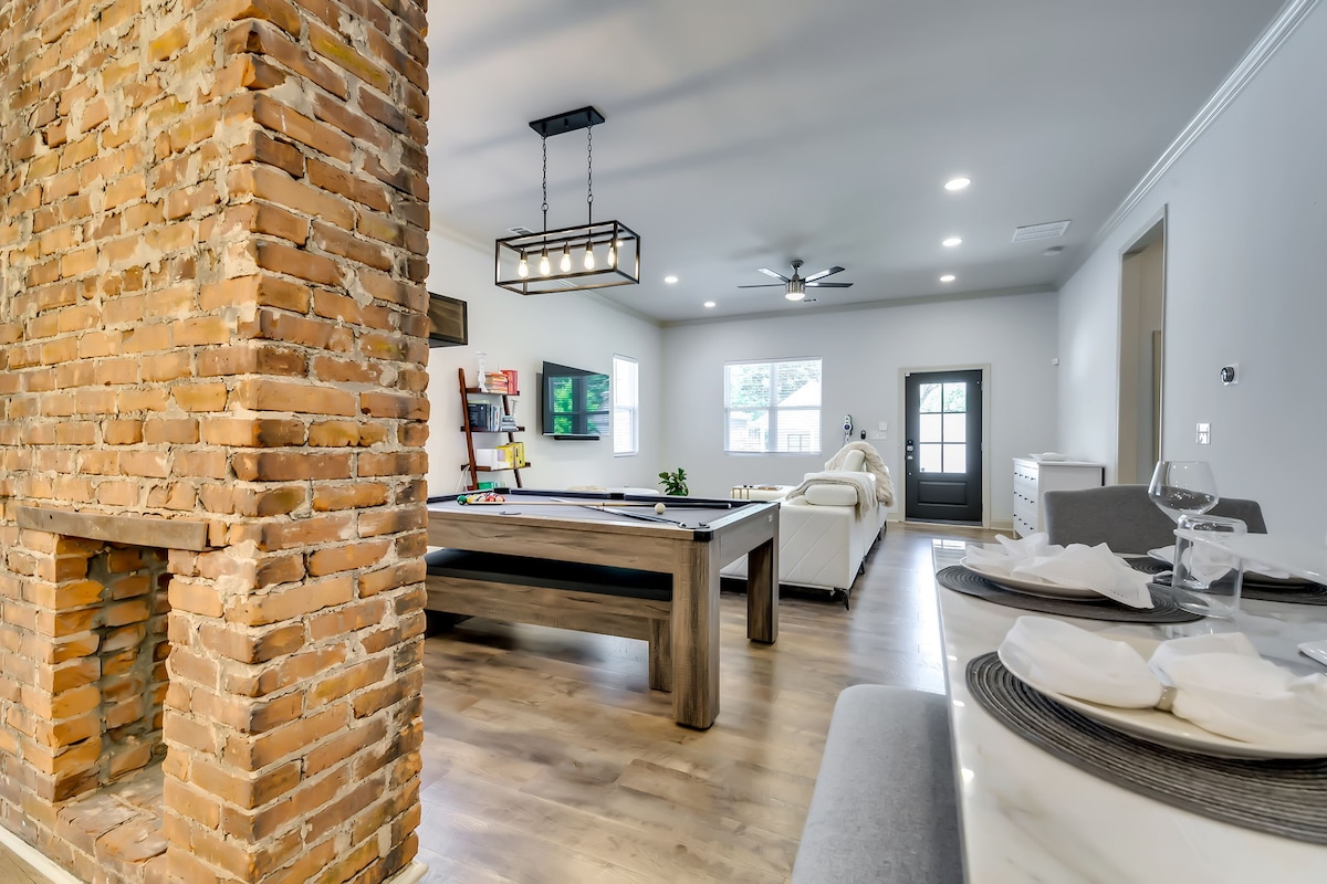 An open floor plan showcases a modern living space featuring a rustic brick accent wall and a view of a dining area. A pool table occupies the center, with comfortable seating visible in the background. Natural light enters through a front door leading to the outdoors.