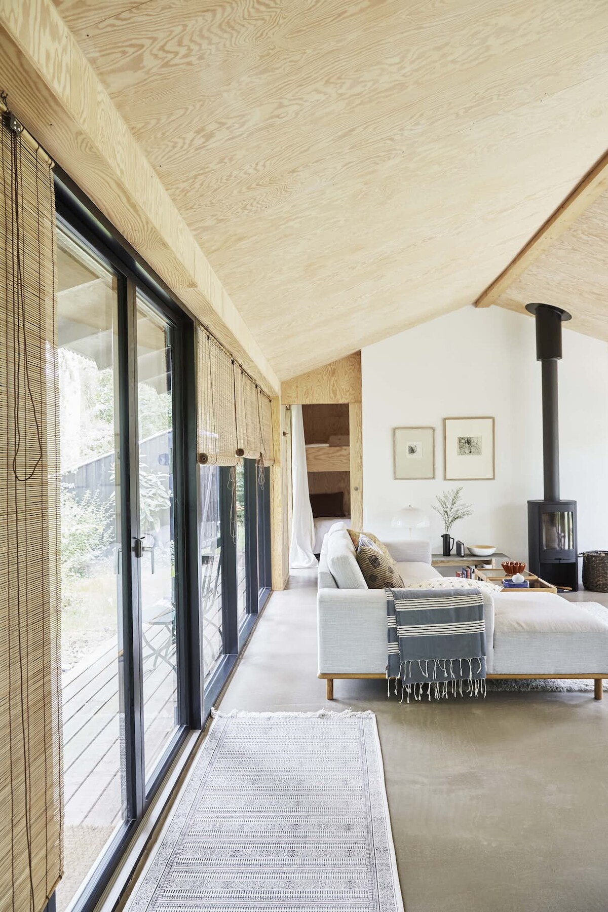 An open living area features a light wooden ceiling and large glass doors that provide access to the outdoor space. A cozy seating arrangement includes a light-colored couch and a wooden coffee table. Decorative elements add character to the tranquil atmosphere.
