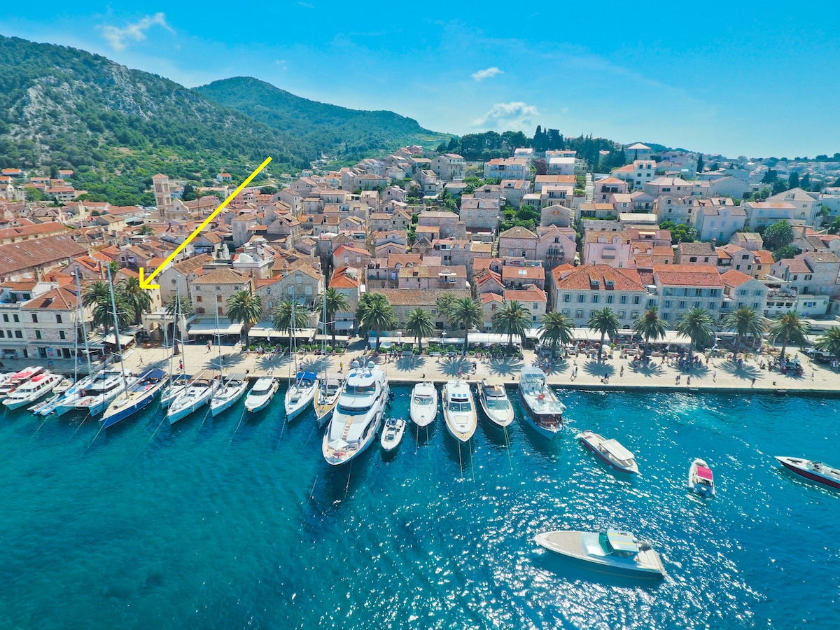 An aerial view captures the vibrant port area, lined with boats docked along the shoreline. Historic buildings with terracotta roofs are visible, set against a backdrop of lush hills under a clear blue sky. The promenade features palm trees, offering a relaxing coastal atmosphere.