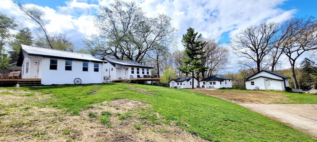 A white, two-story house with large windows sits alongside two smaller buildings, all surrounded by green grass and mature trees. The outdoor space offers a serene setting with an expansive lawn partially sloped towards the structures.