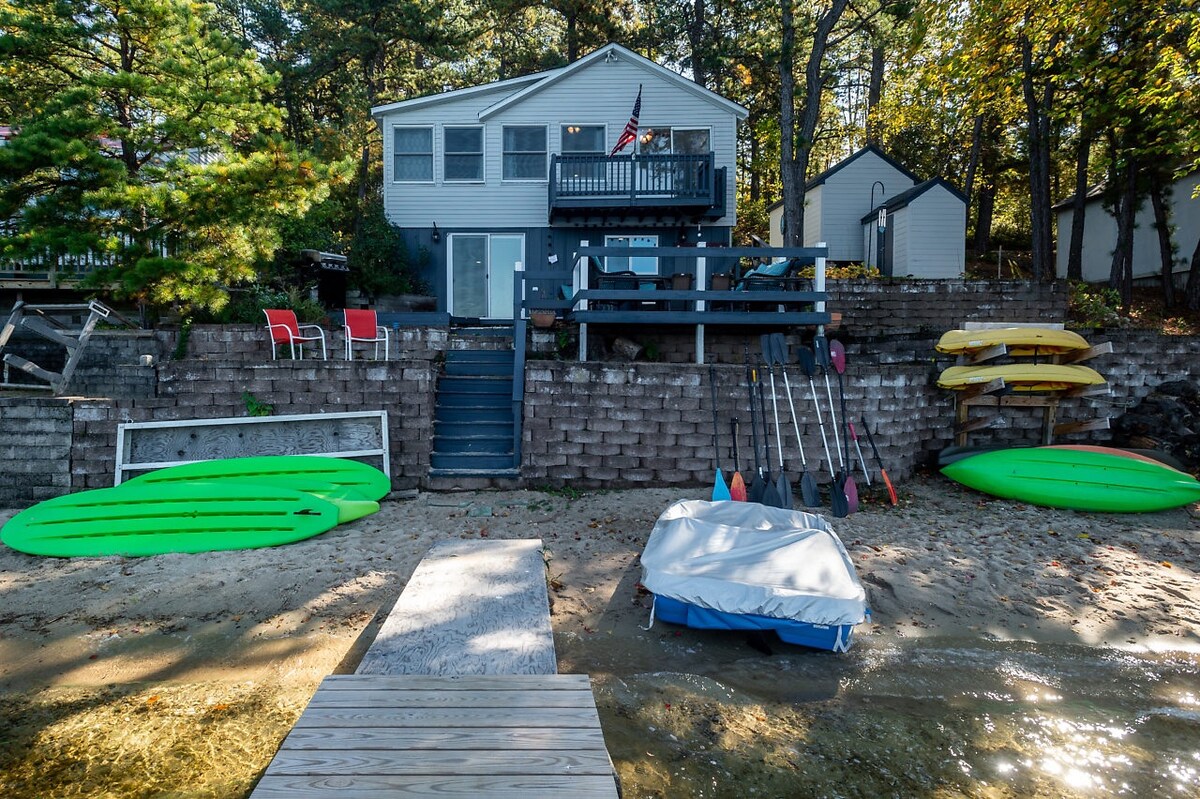A serene lakeside view showcases the two-story lake house with a welcoming deck and stairs leading to the water. Various paddleboards and kayaks rest on the sandy beach, while a small boat cover is visible at the water's edge.