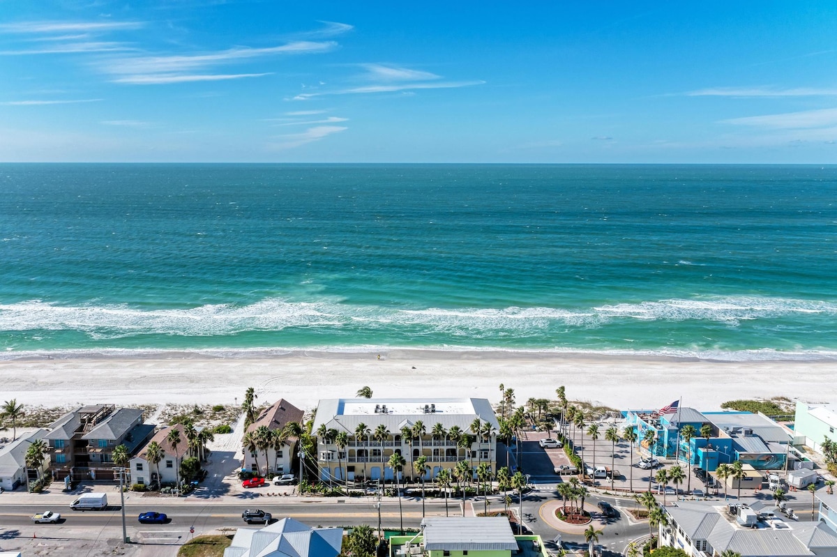 Aerial view captures the expansive Gulf of Mexico, showcasing its vibrant blue waters and gentle waves along the sandy shore. Several beachfront buildings and palm trees are visible, creating a lively coastal atmosphere. The image reflects a clear sky with minimal clouds overhead.