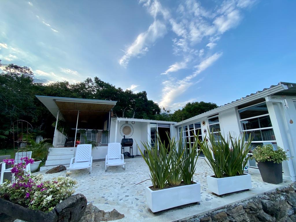 An outdoor patio area features two white lounge chairs facing a natural landscape. Lush green plants and colorful flowers are arranged in planters along the stone pathway. A covered seating area with a barbecue grill is positioned nearby, complemented by a clear sky.