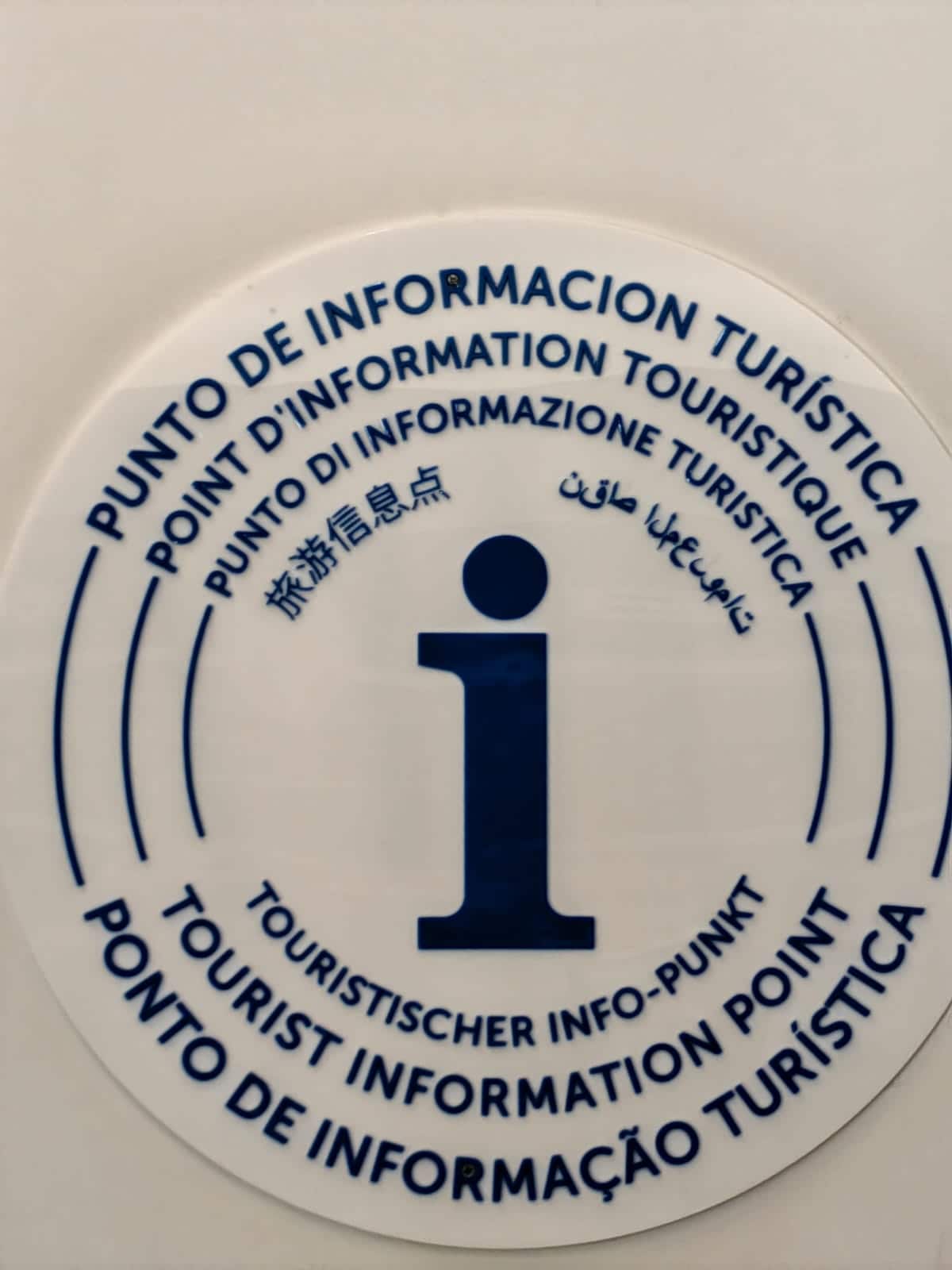 A circular sign features the letter 'i' at its center, representing a tourist information point. The sign displays multilingual text, including Spanish, English, and Arabic, conveying information about local attractions and services for visitors.
