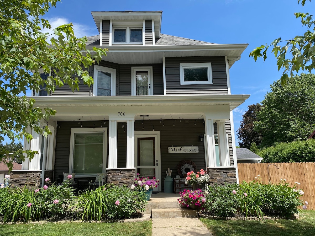 The welcoming entryway of the upper flat features a covered porch adorned with hanging lights and colorful potted plants. The charming exterior includes a mix of dark siding and stone accents, complemented by an inviting front path leading to the main door.