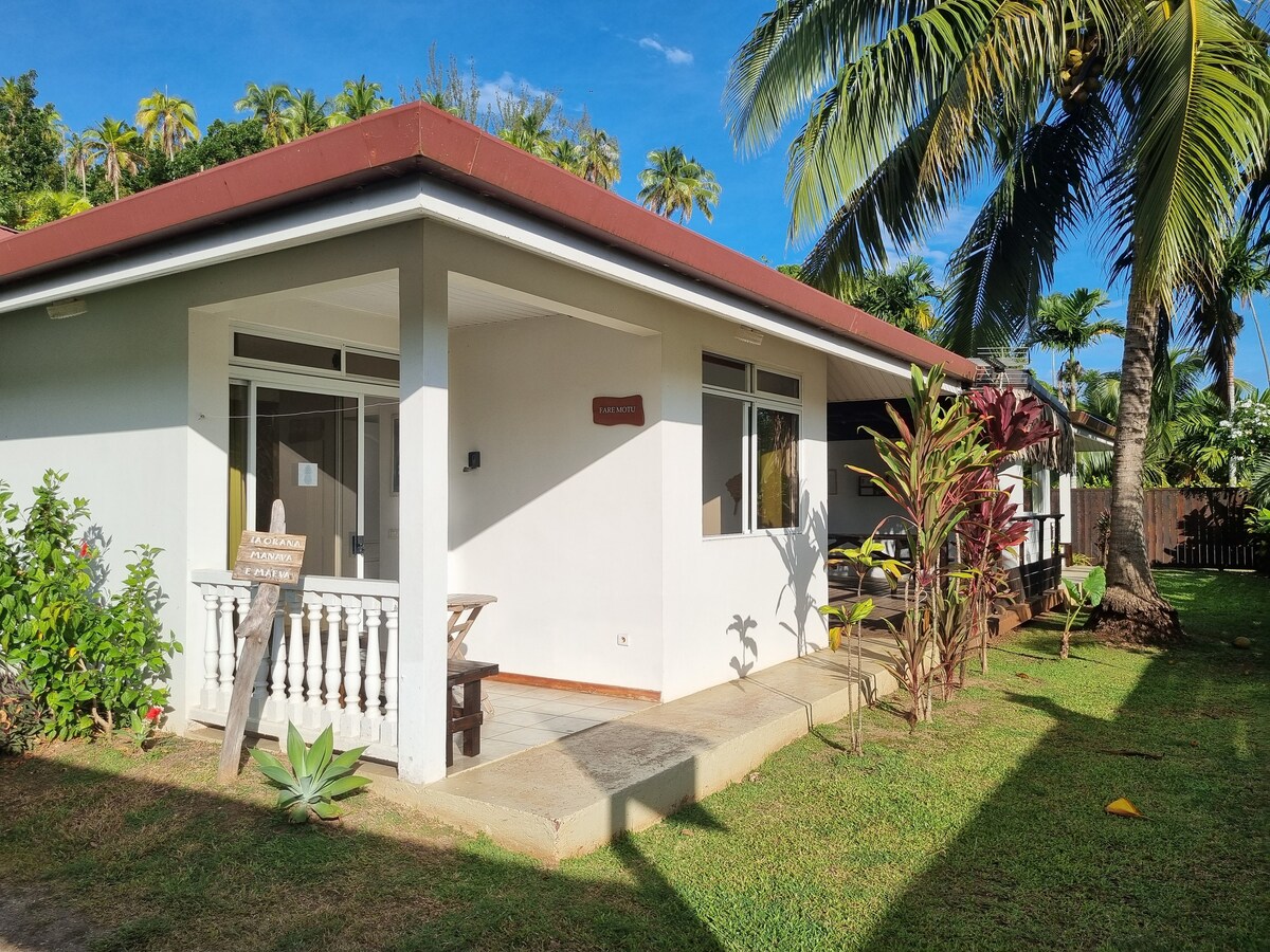 A single-story structure showcases a simple exterior with a red-tiled roof. The entrance is framed by green foliage and palm trees, leading to a charming front patio area. A sign is positioned near the entrance, and a manicured lawn provides a pleasant backdrop.