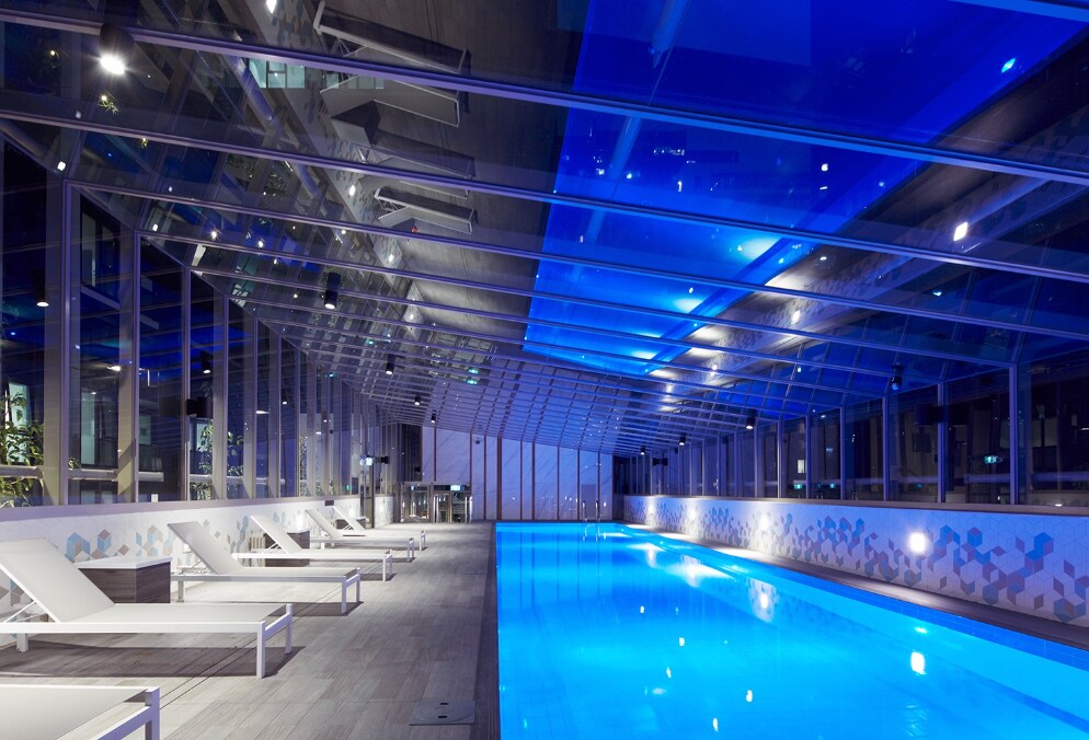 An indoor swimming pool area features a long, illuminated pool surrounded by lounge chairs. The ceiling is equipped with translucent panels that allow natural light to filter through, while blue lighting creates a calming ambiance. The walls are adorned with geometric patterns.