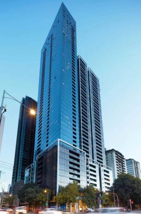 A modern high-rise building is captured against a clear blue sky, showcasing a sleek glass exterior that reflects surrounding urban settings. The structure features multiple levels with visible balconies and is situated among other buildings, presenting a vibrant city atmosphere.
