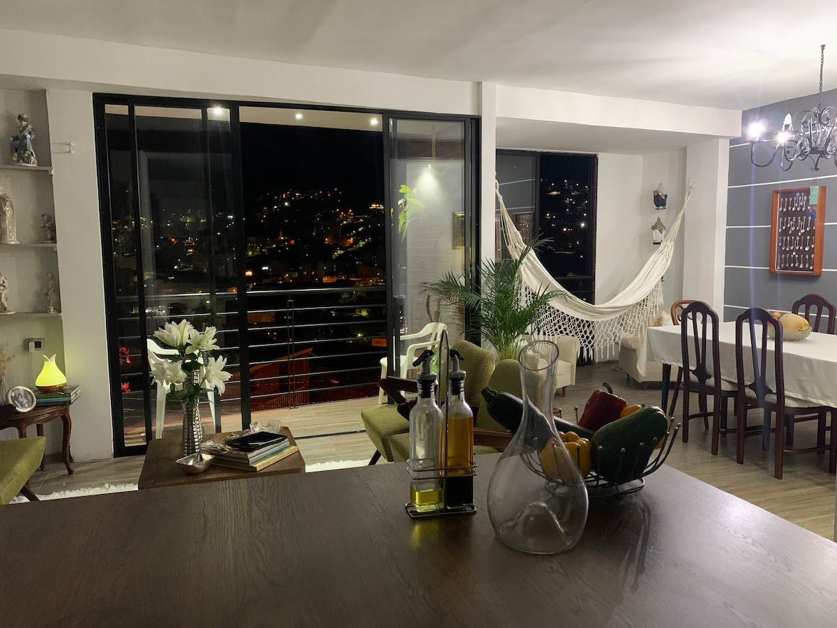 A spacious living area is visible, showcasing large glass doors that open to a balcony with city views. A cozy hammock is suspended, and an elegant dining table is set for four. Decorative items are placed on shelves, and a modern chandelier provides soft lighting.
