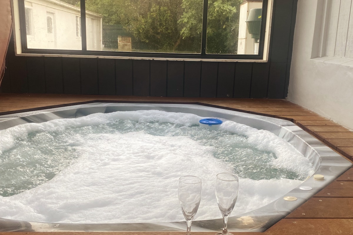 A spacious hot tub is located in an enclosed area with large windows overlooking greenery. Bubbles create a soothing ambiance, accompanied by two champagne glasses placed on the deck nearby, suggesting a relaxing atmosphere.