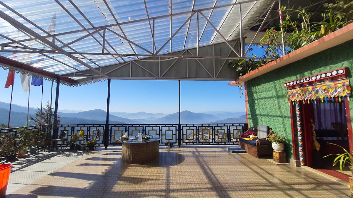 A spacious terrace is covered with a transparent roof, offering panoramic mountain views. Colorful potted plants adorn the area, and a circular seating arrangement is centrally located. The walls display vibrant decorations, enhancing the outdoor atmosphere with a blend of nature and culture.