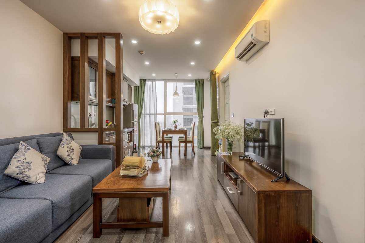 A cozy living room features a comfortable blue sofa, a wooden coffee table, and a TV stand with a flat-screen television. Natural light floods the space through large windows, complemented by a small dining area with a table and chairs. Decorative flowers add a touch of freshness.