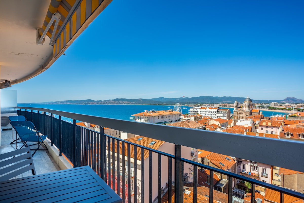 A balcony view features a striking panorama of the bay and cityscape. Bright blue skies complement the red-roofed buildings below, while the ocean glistens in the distance. Two lounge chairs are positioned for relaxation, alongside a small table, enhancing the outdoor seating area.