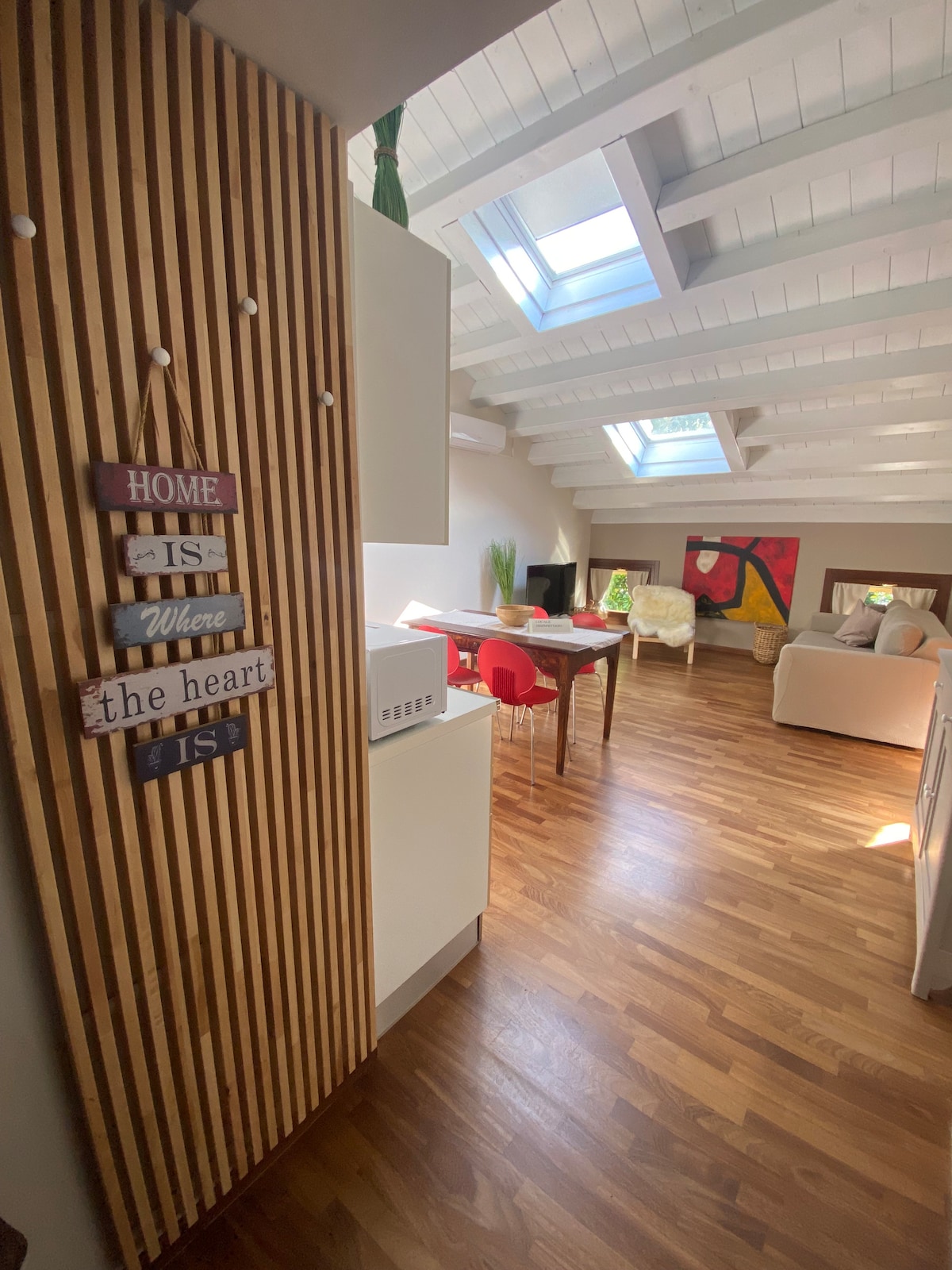 An open space is visible, featuring a modern living area with prominent wooden flooring and slanted white ceilings. The area is accented by skylights allowing natural light. A dining table with red chairs and a cozy seating area are also present, alongside a colorful wall art piece.