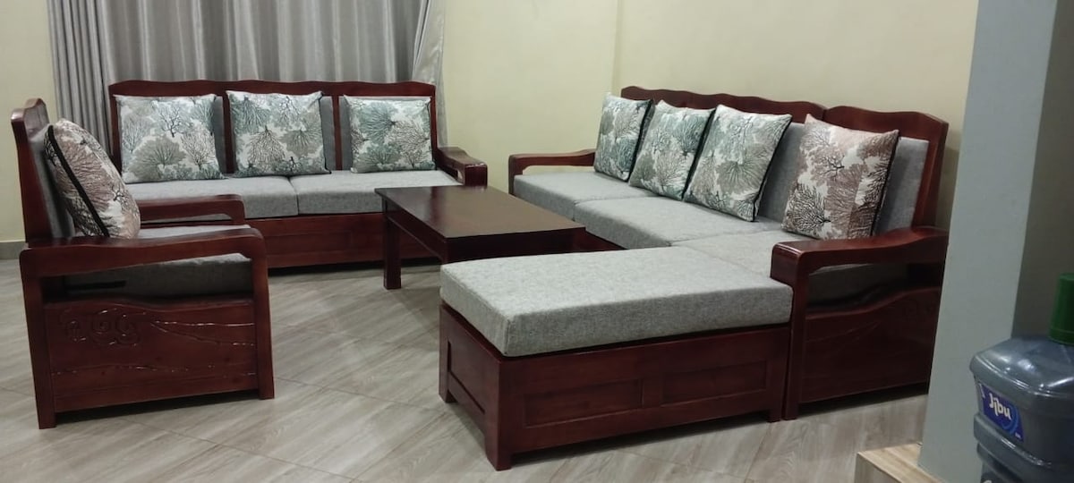 A comfortable seating area is furnished with two wooden sofas, each adorned with patterned cushions. A low coffee table sits in the center, complemented by a cushioned bench that extends the seating arrangement. Light gray walls enhance the relaxed ambiance.
