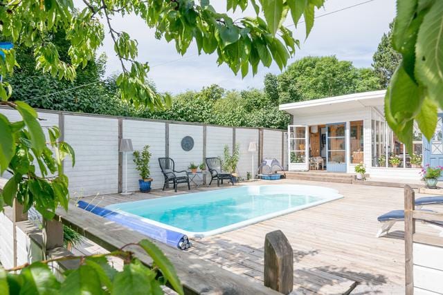A serene backyard features a small, inviting pool surrounded by a wooden deck. Lounge chairs are positioned nearby, with vibrant greenery providing shade. The exterior of a light-colored house is visible, framed by open doors and windows that enhance the spacious feel.