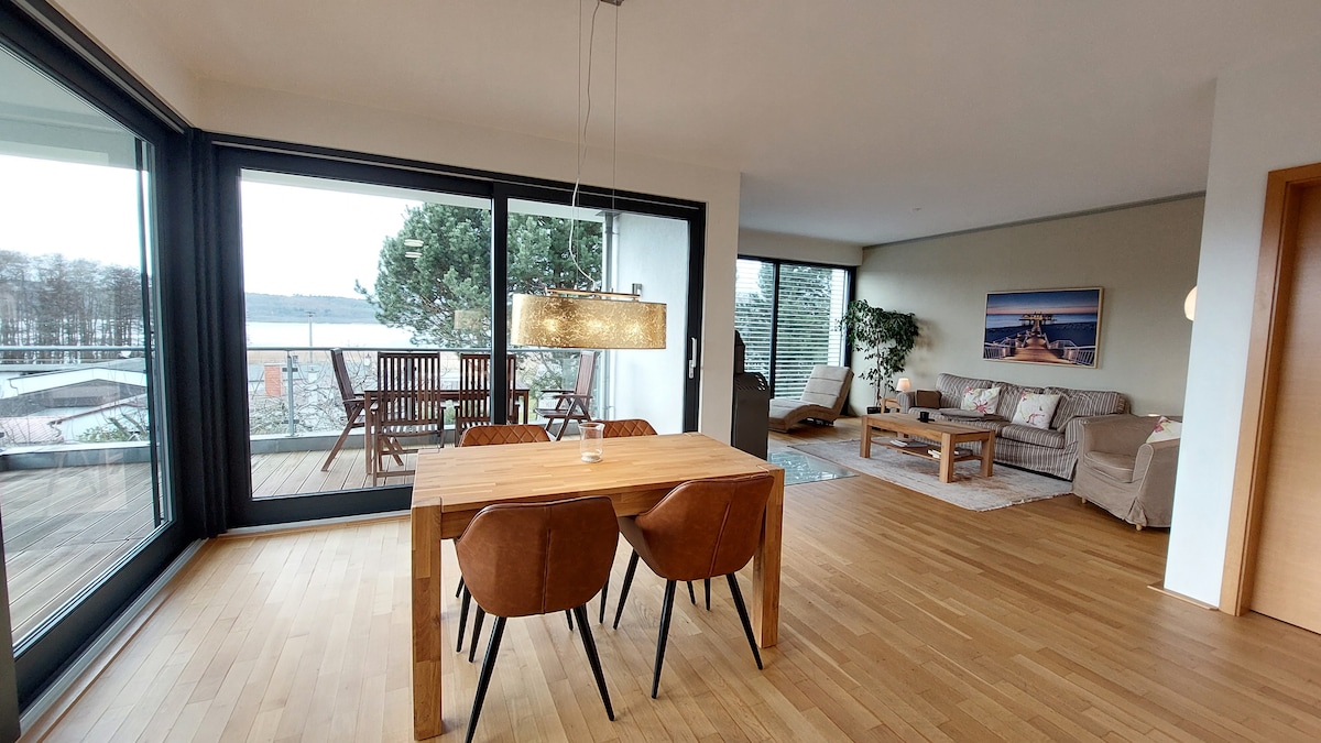 A spacious living area features a modern dining table made of light wood, accompanied by four brown chairs. Large glass doors offer views of a balcony and the surrounding landscape, allowing natural light to fill the room. Cozy seating arrangements and decorative elements create a welcoming ambiance.