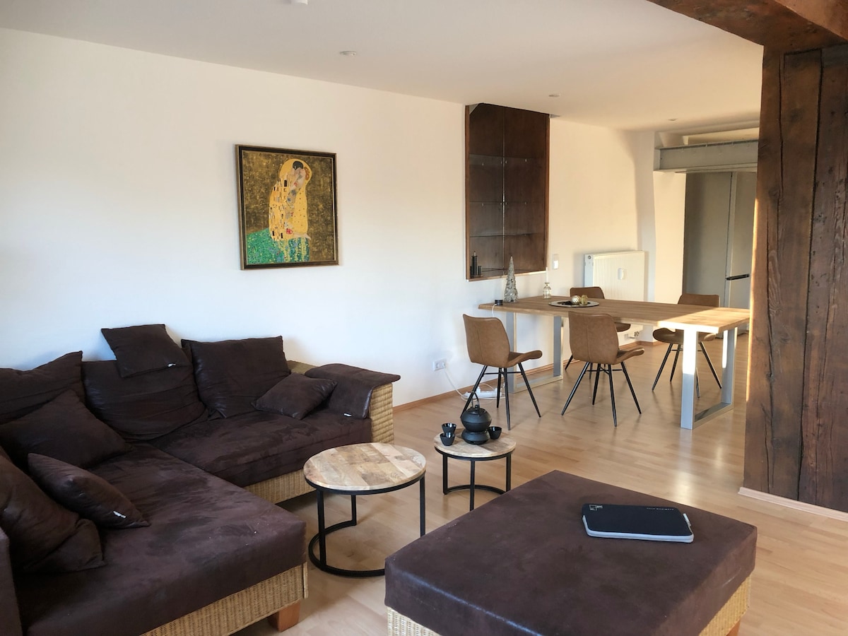 A comfortable living area is depicted with a brown sectional sofa and a matching ottoman. Two circular wooden tables occupy the space, alongside a dining table set for four. A vibrant piece of wall art is displayed prominently, enhancing the modern atmosphere.