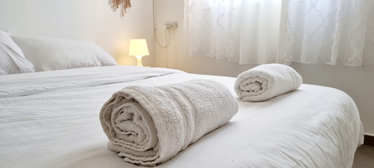 A spacious bed is covered with crisp white linens, complemented by neatly rolled towels positioned on top. Soft light from a nearby lamp illuminates the room, which is adorned with sheer curtains allowing gentle sunlight to filter through.