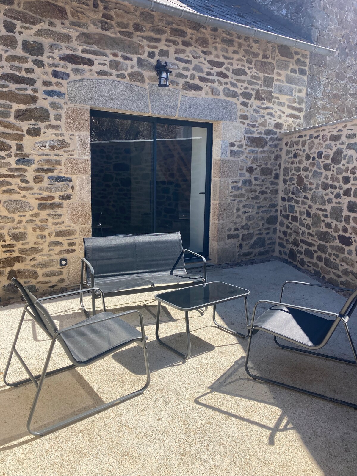 An outdoor patio space features a stone wall backdrop and a modern glass door. Four minimalist seating chairs surround a glass-top table on a sandy surface, providing a comfortable area for relaxation. Natural light enhances the serene atmosphere.