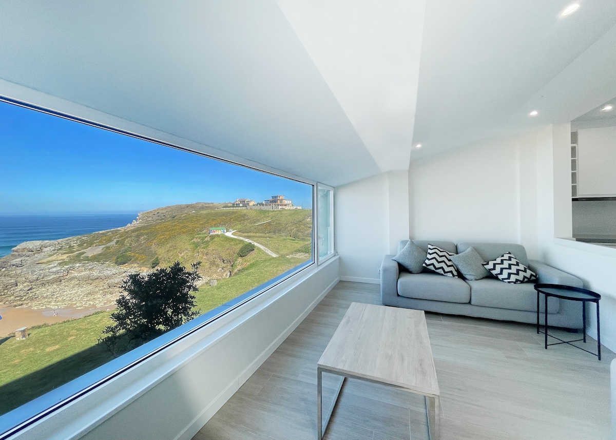 A spacious living area features a large window, offering expansive views of the coastline and the beach. A light-colored sofa, adorned with patterned cushions, is complemented by a wooden coffee table. Natural light enhances the bright, airy atmosphere of the room.