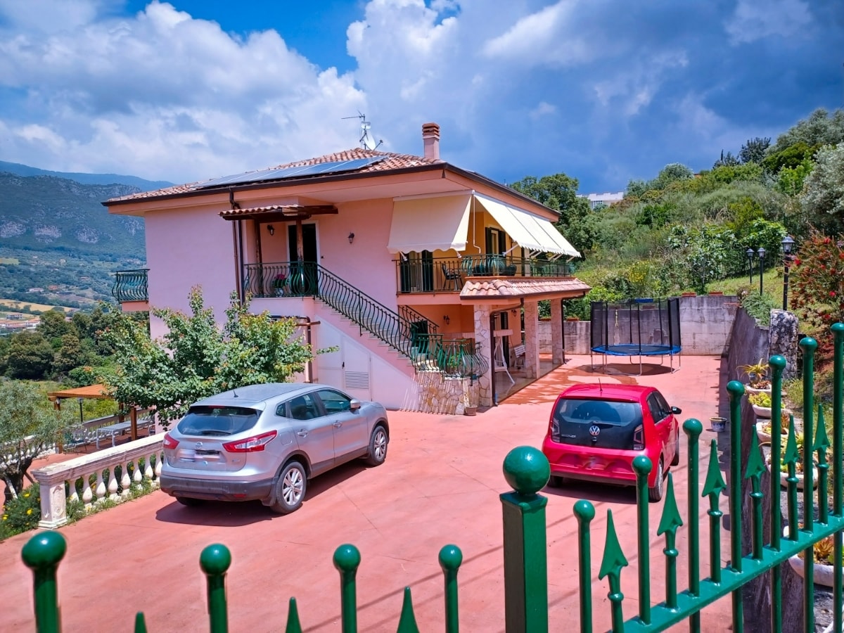A two-story house is situated on a hillside, surrounded by greenery. A paved driveway provides access to the property, which features two parked vehicles. A trampoline can be seen nearby, with rolling hills and clouds in the background.