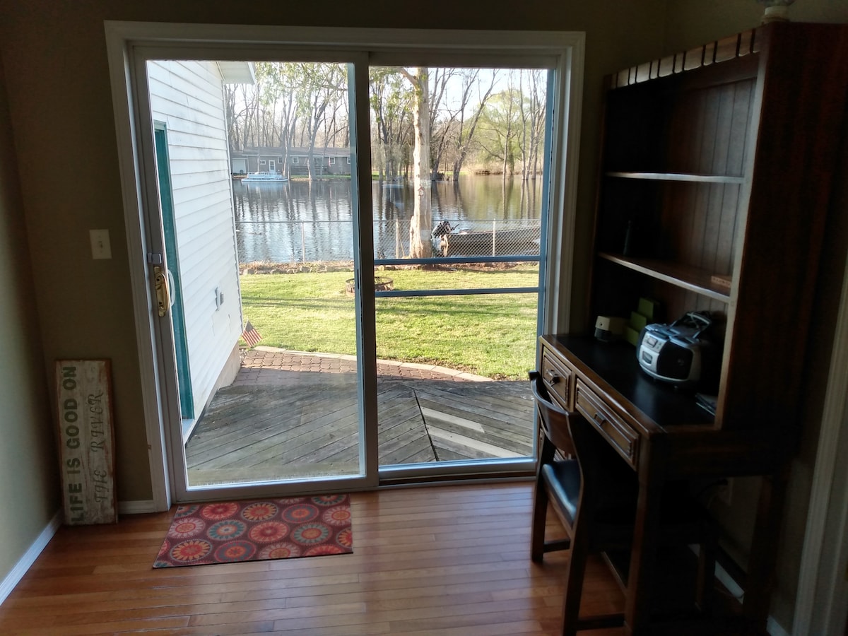 A bright room featuring a large sliding glass door that opens to a scenic view of the river and greenery outside. A wooden desk with a chair is positioned nearby, providing a functional workspace. The polished wooden floor complements the inviting atmosphere.