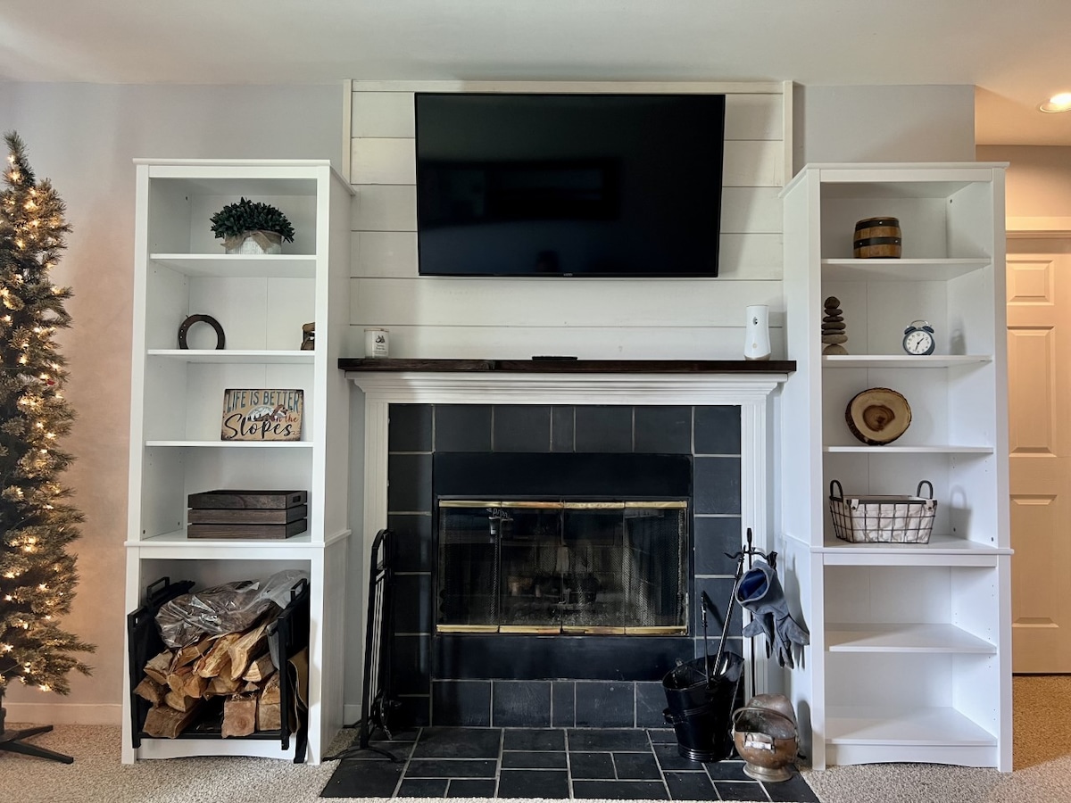 The living area features a wood-burning fireplace framed by a black slate surround. Tall white bookshelves on either side display various decorative items. A flat-screen TV is mounted above the mantel, with a stack of firewood neatly stored beside the fireplace.