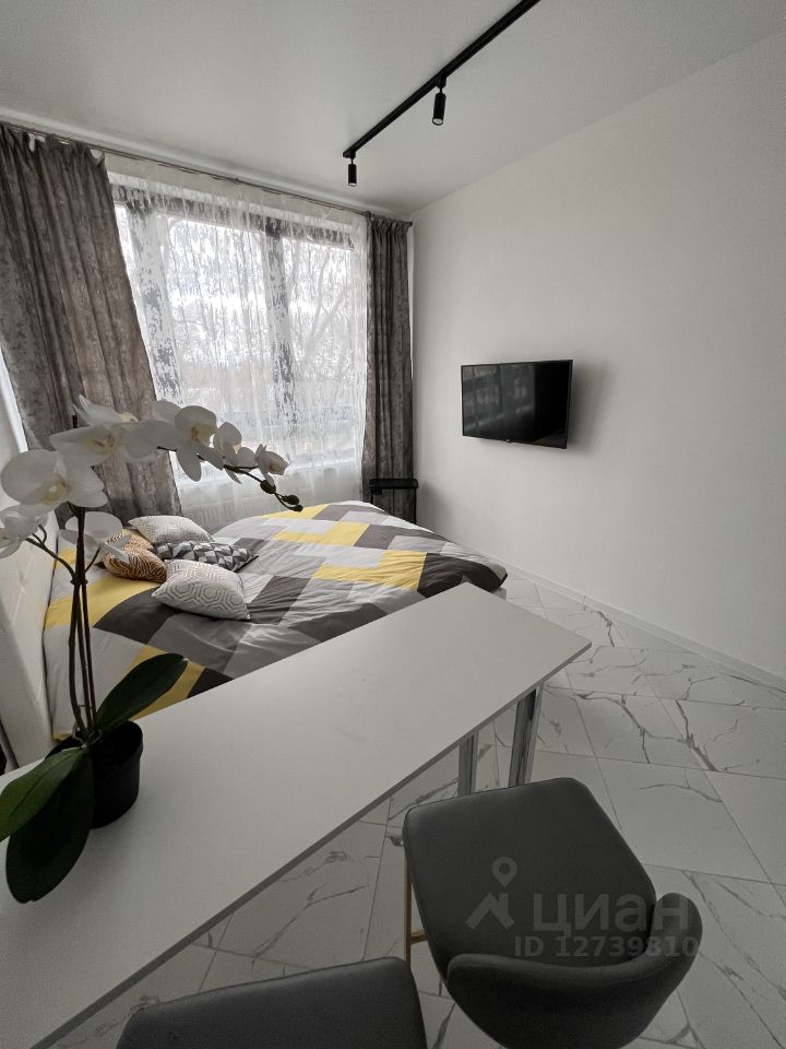 A cozy sleeping area features a spacious bed dressed in a colorful quilt. Natural light filters through sheer curtains, illuminating the room. A minimalist table sits nearby, while a television is mounted on the wall opposite the bed, enhancing the modern decor.