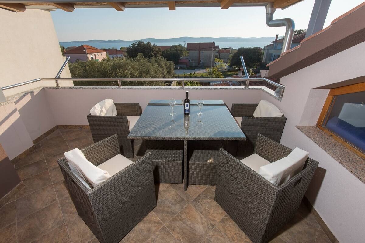 A balcony seating area is featured, showcasing a glass-top table surrounded by four wicker chairs with cushions. A scenic view of the surrounding landscape is visible in the background, while the flooring is composed of large, patterned tiles.